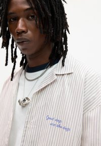 Camicia a righe bianca con linee viola. Presenta testo ricamato "I giorni belli sono giorni lenti" e collane sovrapposte.