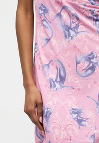 Κοντινό πλάνο ενός χεριού που ακουμπά σε ροζ ύφασμα με μοτίβο μοβ γοργόνας και διακοσμητικές σπείρες.