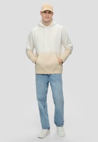 Sudadera con degradado beige, bolsillo frontal, cordones, mangas largas, combinada con jeans de un azul claro y zapatillas blancas. Gorro en color a juego.