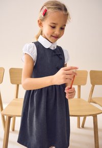 Grå denimkjole med en rynket nederdel, der har en hvid skjorte med krave under og to dekorative sløjfer. Træstole i baggrunden.