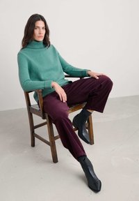 Tirkizni puli, rebraste manžete, u paru s tamno burgundskim pamučnim hlačama. Model nosi crne čizme do članaka, sjedi na drvenoj stolici.