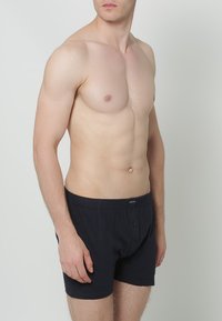 Zwarte katoenen boxers met een knoopsluiting, voorzien van een zachte tailleband en zonder zichtbare patronen, gedragen door een mannelijke model voor een neutrale achtergrond.