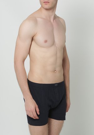 Zwarte katoenen boxers met een knoopsluiting, voorzien van een zachte tailleband en zonder zichtbare patronen, gedragen door een mannelijke model voor een neutrale achtergrond.