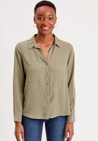 Blouse olive verte à manches longues avec col, fermeture à boutons et deux poches poitrine. Tissu lisse avec une coupe décontractée et un ourlet arrondi.