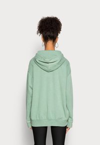 Person med lockigt hår i en hög knut, bärande silverörhängen med ring och en lös, salviafärgad huvtröja, vänd bort från kameran mot en vit bakgrund.