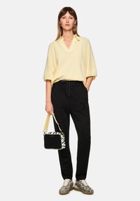 Gelbes übergroßes Strick-Poloshirt mit V-Ausschnitt, schwarze Jogginghosen mit einem dezenten Zebramuster und schwarz-weiß gemusterte Schuhe.