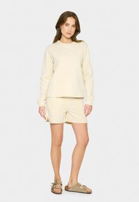 Sudadera de algodón beige con logo bordado, combinada con pantalones cortos beige con aberturas laterales. Llevado con sandalias marrón claro. Diseño simple.