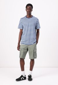 Stribet blå og hvid t-shirt, grønne cargo shorts med lommer, sorte sneakers og hvide sokker, stående mod en ensfarvet baggrund.