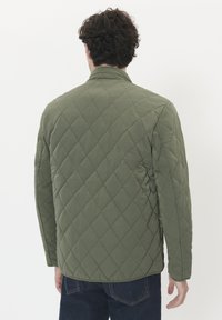 Mann trägt eine grüne Steppjacke und dunkelblaue Jeans, steht mit dem Rücken zu einem schlichten weißen Hintergrund.