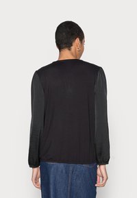 Svart topp med långa, glänsande svarta ärmar och en mjuk textur. Har en rund halsringning och rynkade manschetter, i kombination med en jeanskjol.