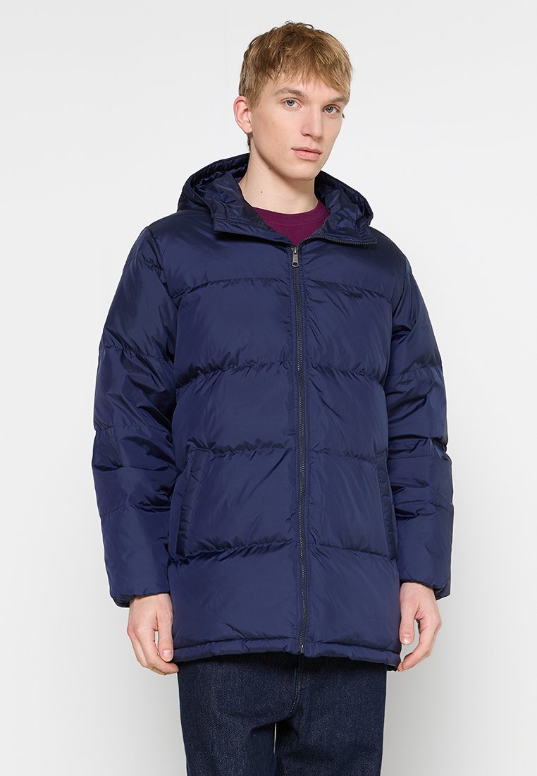 Doudoune bleu marine avec zip intégral, capuche et deux poches avant. Présente un design matelassé et une texture lisse et brillante.