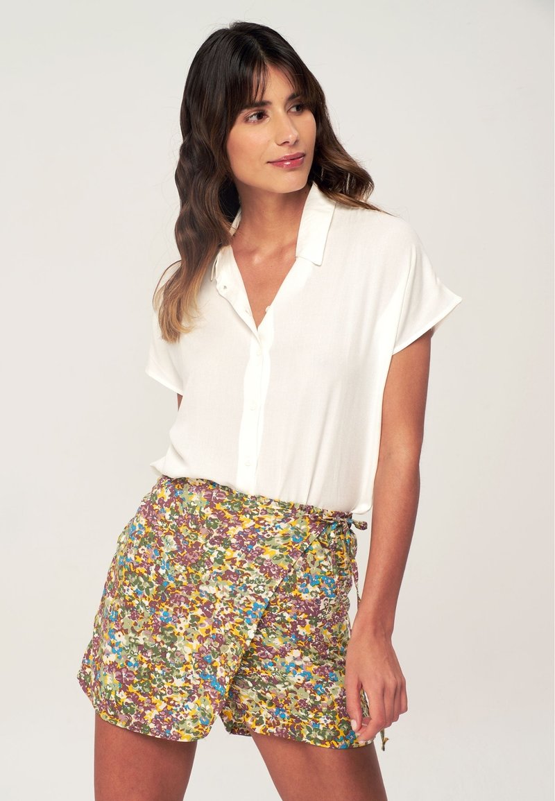 Camicetta bianca a maniche corte con bottoni abbinata a pantaloni corti che presentano un motivo floreale colorato, con vita legata e vestibilità ampia.