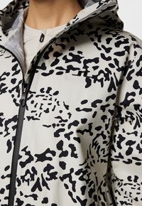 Lekka kurtka w kolorze szarym z czarnym abstrakcyjnym wzorem, z zamkiem z przodu, regulowanym kapturem i małym logo na piersi.