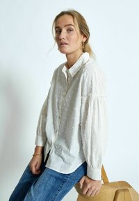 Witte button-up blouse met een gestructureerd stippenpatroon, met een kraag en pofmouwen. Gecombineerd met blauwe denim jeans en houten accenten.