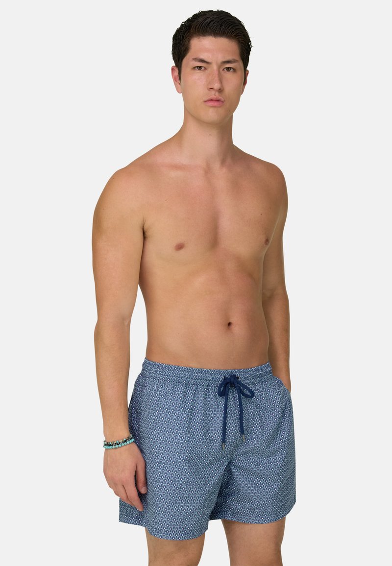 Homme torse nu portant un short de bain bleu à motifs avec cordon, debout avec la main gauche dans la poche et portant un bracelet de perles au poignet droit.