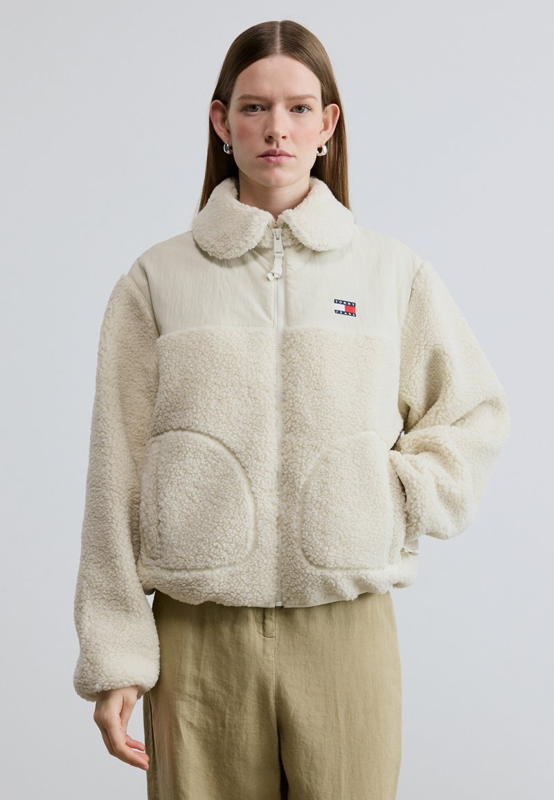Μπουφάν σε κρεμ απόχρωση με απαλή υφή ψεύτικου shearling, φερμουάρ μπροστά, πλαϊνές τσέπες και στρογγυλεμένο γιακά. Διαθέτει λογότυπο στο στήθος.