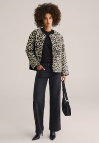 Leopardmönstrad vadderad jacka med rundad halsringning, svart långärmad tröja, svarta vida jeans och svarta högklackade stövlar, hållande en väska.