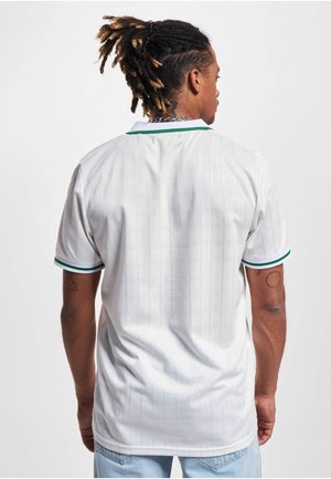 Mand med dreadlocks iført en hvid kortærmet poloshirt med grøn kant, set bagfra, og lyseblå jeans mod en ensartet hvid baggrund.