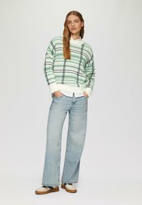 Hellgrüner gestreifter Pullover mit schwarz-weißen Akzenten, kombiniert mit hellblauen weit geschnittenen Jeans und braunen sowie weißen Schuhen.