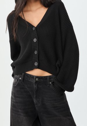 Frau trägt eine schwarze, grob gestrickte, geknöpfte Strickjacke mit Ballonärmeln und hoch taillierte, ausgewaschene schwarze Jeans.