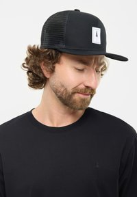Gorra negra con visera plana y lados de malla que presenta un parche rectangular blanco con un gráfico de un árbol. Se lleva con una camiseta negra de cuello redondo.