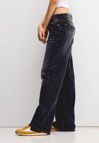 Person som bär lösa svarta jeans, en vit croppad topp och gula och vita sneakers, står mot en enkel vit bakgrund.