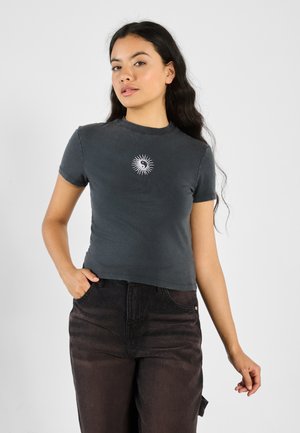 Grå kortermet t-skjorte laget av bomull, med et hvitt solstråledesign i midten. Kombinert med mørke denimbukser.