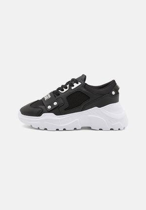 Sneaker chunky bianco e nero con pannello in mesh, suola spessa e strutturata, chiusura frontale con lacci e cinturino decorativo con testo su sfondo grigio.
