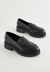 Mocassins en cuir noir avec un bout rond, présentant un détail de bande à l'avant et une semelle en caoutchouc épaisse pour plus de hauteur et d'adhérence.