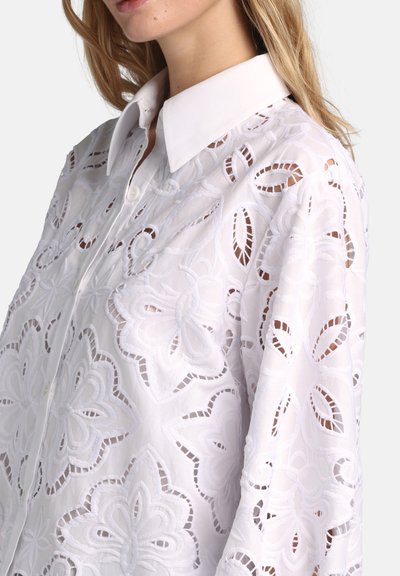 Frau trägt ein weißes Hemd mit großen floralen Cutout-Stickereien und spitzem Kragen, zeigt Oberkörper und Hals.