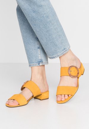 Sandales en daim jaune avec un talon épais, un design ouvert à l'avant et des bretelles larges. Une des bretelles présente un grand accent de boucle circulaire.