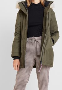 Parka imbottita verde oliva con cappuccio foderato in pelliccia, chiusura con zip e polsini a coste neri, abbinata a pantaloni a quadri con cintura a cravatta.