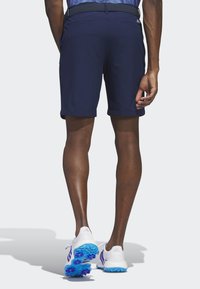 Tamsiai mėlyni šortai su moderniu dizainu, matomomis nugarinėmis kišenėmis, derinami su baltais ir mėlynais sportiniais bateliais, kurių pėdos apačia turi ryškų protektorių.