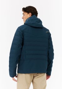 Námornícka prešívaná bunda s kapucňou, so hladkou textúrou a logom na pleci. Skombinovaná so svetlými nohavicami.