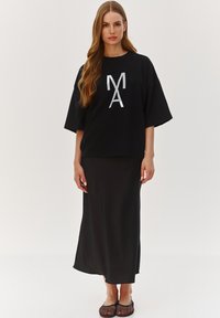 Czarny, oversize t-shirt z odblaskowym srebrnym logo, w połączeniu z długą czarną spódnicą. Noszony z czarnymi, siateczkowymi butami na płaskiej podeszwie. Gładka tekstura materiału.