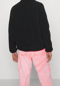 Černá fleecová bunda s vysokým límcem, zaobleným spodním lemem a raglánovými rukávy, zkombinovaná se světle růžovými džíny se viditelným švem.