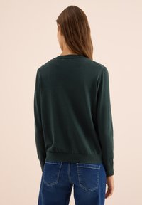 Frau mit langen braunen Haaren, die einen dunkelgrünen Pullover und blaue Jeans trägt, von hinten vor einem beigen Hintergrund gezeigt.