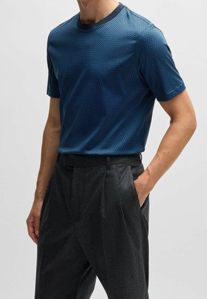 Mann trägt ein tailliertes, kurzärmeliges blau gemustertes Hemd, in hoch taillierte dunkelgraue plissierte Hose gesteckt, eine Hand in der Tasche.