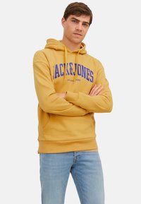 Gelbe Kapuzenjacke mit einer vorderen Kängurutasche und blauer "JACK & JONES"-Logo. Enganliegendes Design mit einer Kordelzugkapuze, kombiniert mit hellblauen Jeans.