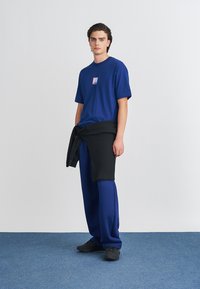 Tumši zils T-krekls ar mazu grafiku, apvienots ar brīvi pieguļošām tumši zilām biksēm. Melna džemperis sasieta ap jostasvietu piešķir tekstūru. Melni sporta apavi.