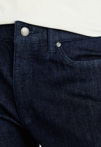 Dark denim jeans featuring a buttoned waist with a metal Lee logo, subtle texture, and a classic five-pocket design.