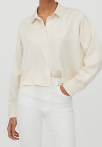 Camicia di colore crema con bottoni, colletto, vestibilità rilassata, maniche lunghe e texture liscia, abbinata a jeans bianchi a vita alta.