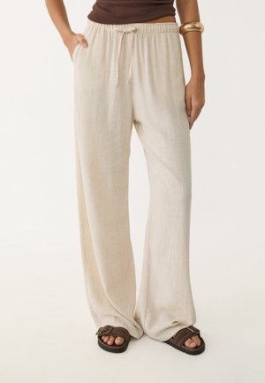 Persoon die losse beige linnen broek, bruine sandalen en een bruin topje draagt, met één hand in de zak en een gouden armband aan de arm.