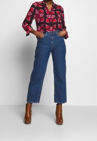 Blouse à motifs rouges et noirs avec un col, associée à un jean en denim bleu taille haute et des bottines brunes. Texture lisse, coupe cintrée.
