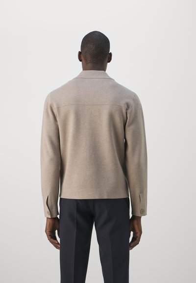 Bež kudumärgi pullover kaelusega, pikkade varrukatega ja nööp detailidega mansettides, koos tumedate pükstega. Sile tekstuur ja keha lähedane lõige.
