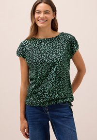 Žena se usmívá, má na sobě černý top s krátkým rukávem s zelenými abstraktními skvrnami a modré džíny, stojí na béžovém pozadí.