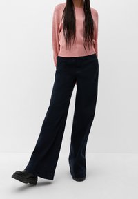 Personne avec de longs cheveux tressés portant un pull rose, un pantalon large bleu marine et des chaussures à plateforme noires sur fond blanc.