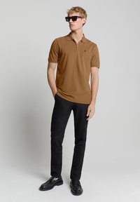 Bruine poloshirt met ritskraag, korte mouwen, logodetail. Gecombineerd met zwarte broek en zwarte instappers, model draagt een zonnebril.