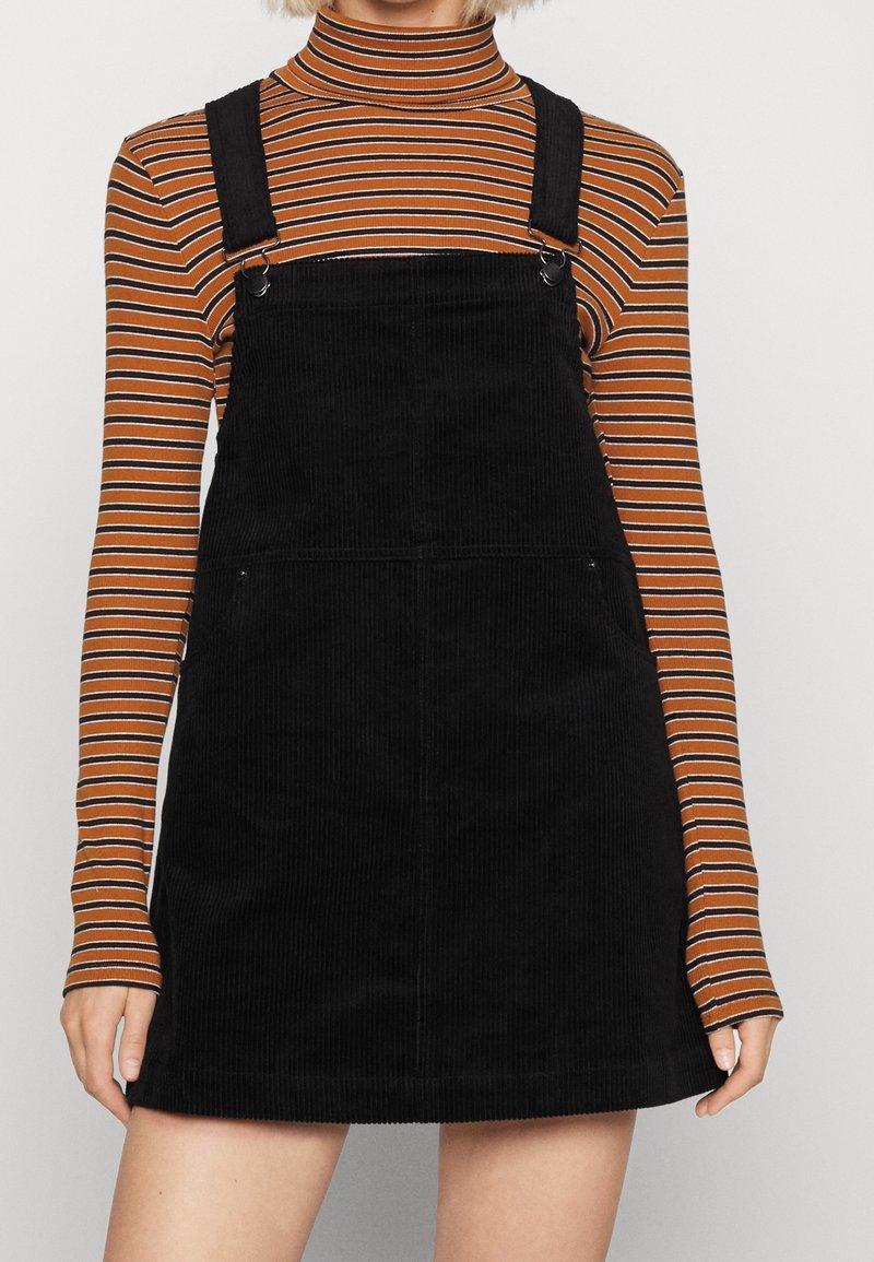 Personne portant une robe salopette en velours côtelé noire sur un top à col roulé rayé marron et noir à manches longues, debout devant un fond uni.