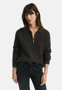 Brauner strukturierter Pullover mit einem Reißverschlusskragen und gerippten Bündchen, kombiniert mit schwarzen Jeans. Der Pullover hat ein Wabenmuster.